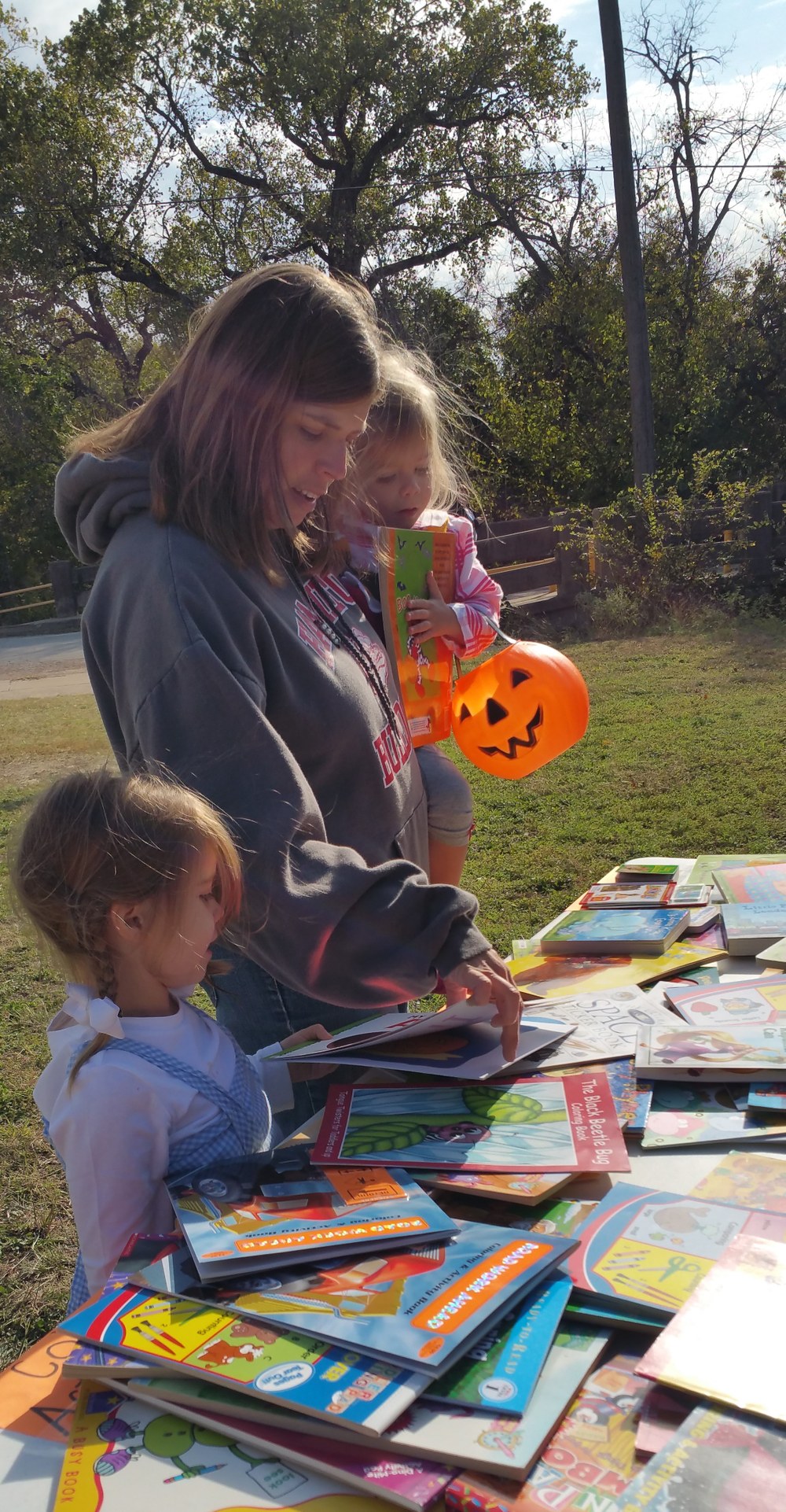 A mother holding a young child in a Halloween costume accompanied by another young child in costume picks a free book from a table with many choices.